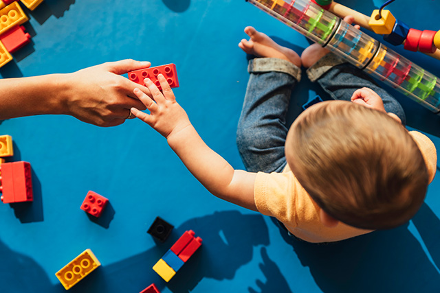 a small child reaching for a block from the hand of an adult